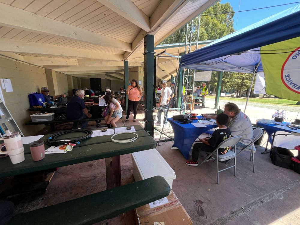 Wide-angle view of two tables in the park shelter with plenty of radio gear on top of each. Young kids are learning to call CQ and make QSOs for Field Day. 