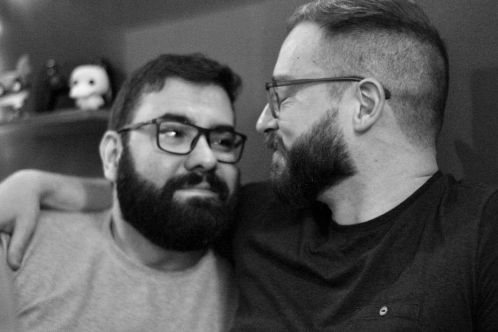 Foto em preto e branco com dois homens de barba e óculos se amando