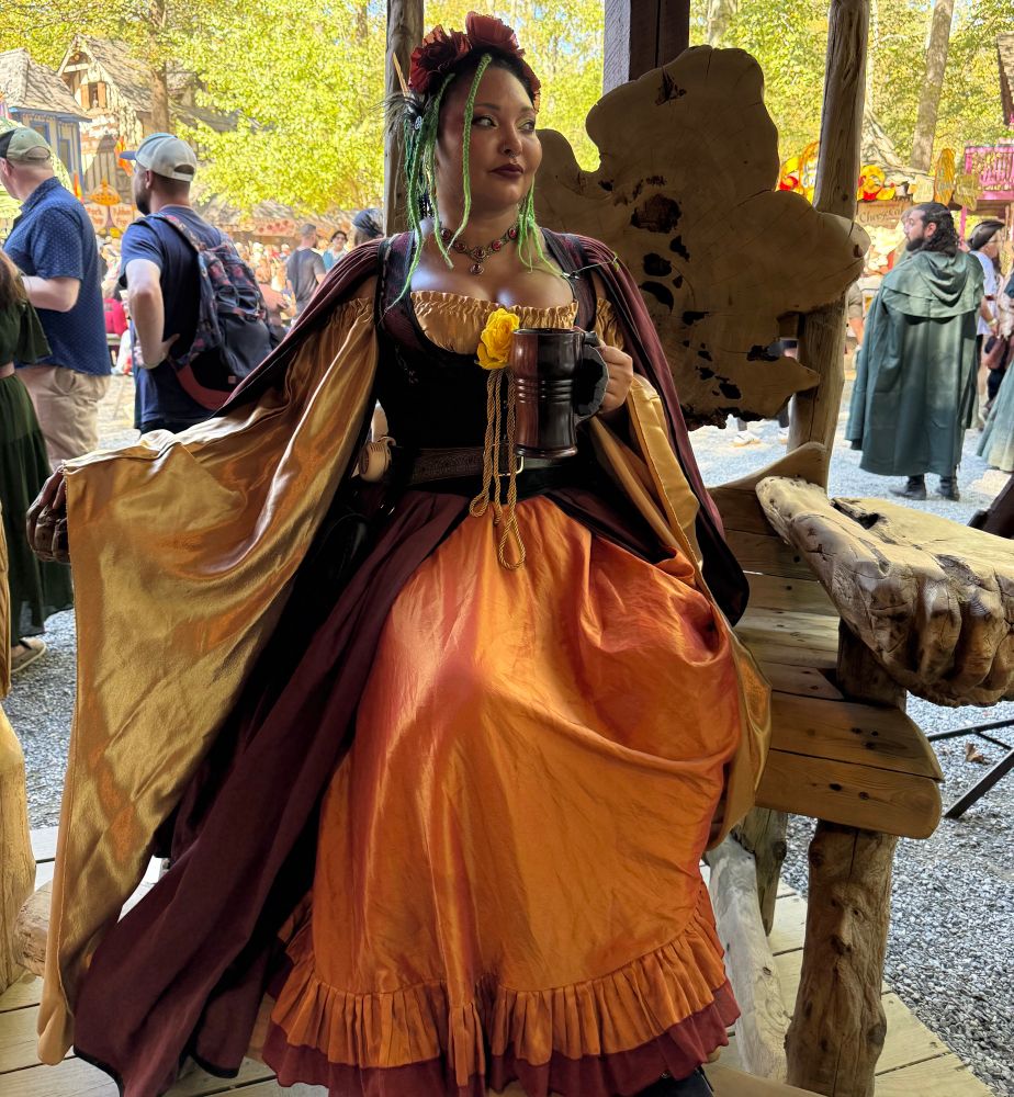 Harvest goddess for rennfest. Sitting in a giant chair holding a wooden mug, dressed in layers of gold, orange, and red, with a flower crown. 