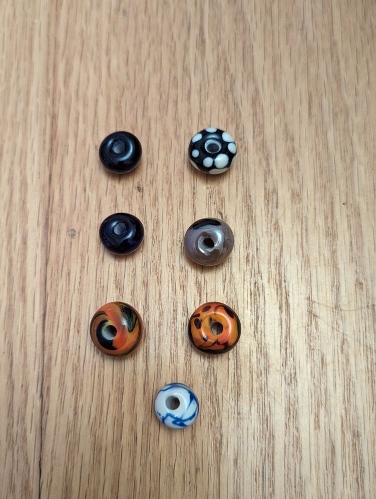 Seven hand-made glass beads, each about a half-inch across. Two dark blue. Two orange with blue swirls. One white with blue swirls. One purple with black swirls. One black with white dots.