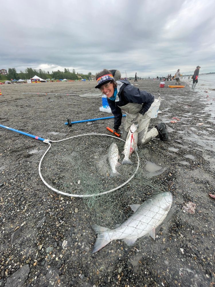 A white woman kneeling with 3 red salmon in a net. One of them is dripping blood, the other two still laying on the ground