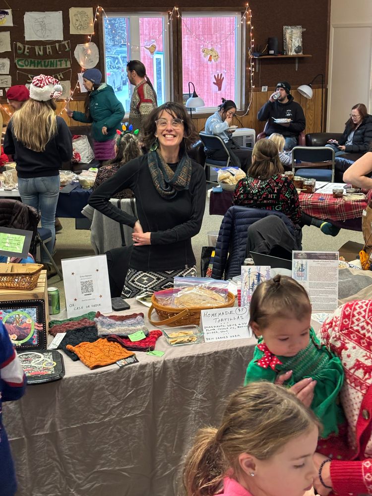 A busy photo of a holiday bazaar, with me smiling at the photographer from behind a kind of themeless sales table, where I’m selling crafts, homemade tortillas, and books 