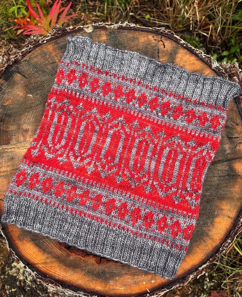 A gray and black knit cowl sitting on a spruce tree stump. The cowl is a 2x2 ribbing on the ends, and colorwork that might be thought to be an abstract representation of mountain peaks, sort of a Scandinavian feel to the aesthetic 