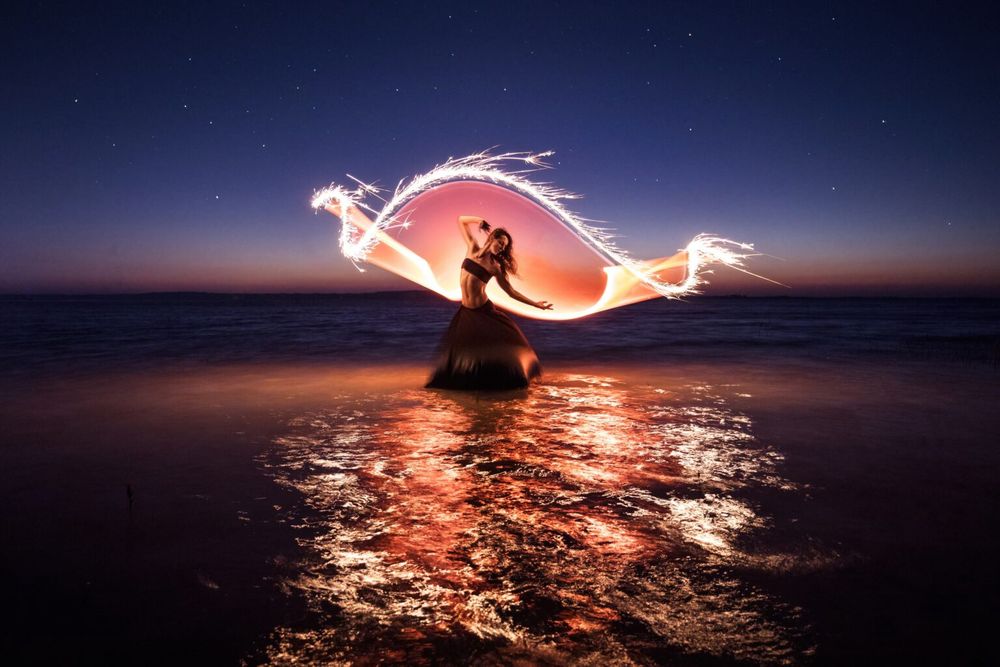 A photo by Eric Paré. It's twilight and there is a woman striking a pose, behind her is a twisted ribbon of light and sparks, which is reflected in the rippled water she's standing in.