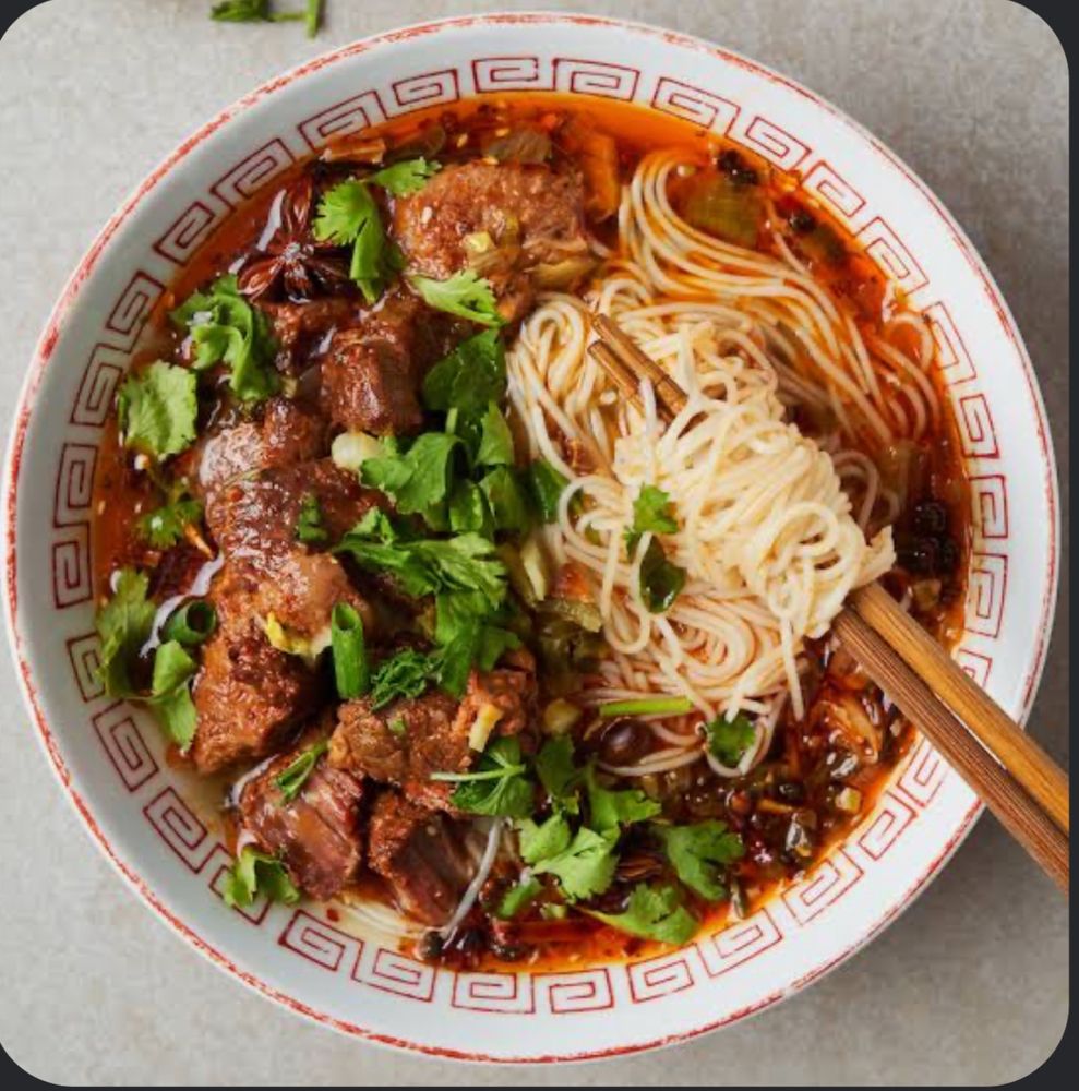 A bowl of soup with thin noodles, chunks of beef, cilantro, and a red spicy broth. Chopsticks have slightly lifted the noodles to show them off. 