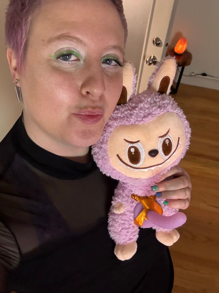 Star wears a black mesh long sleeve shirt and smiles at the light purple plushie with a smiling cute but demonic face. The plushie has a light purple dildo strapped onto its crotch. 