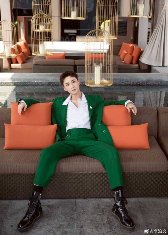 Li Bowen in the GREEN SUIT and black high-top shiny shoes sitting on a brown couch with orange pillows manspreading. In the background weird little bird cages with candles in them LITTER every surface. 