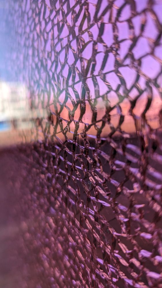 ​Dies ist eine Nahaufnahme durch eine Glasscheibe, die in viele kleine, unregelmäßige Stücke zerbrochen (oder gesplittert) ist, aber noch in ihrem Rahmen hält (Sicherheitsglas oder Verbundglas).
​Details und Stimmung
​Vordergrund (Textur): Die Oberfläche des Glases ist von einem dichten Netz aus Rissen durchzogen, die wie kleine, miteinander verbundene Mosaikstücke aussehen.
​Farbgebung: Das gesamte Bild hat einen starken, durch das Glas gefilterten violetten oder magentafarbenen Stich, der die Lichtstimmung dominiert.
​Hintergrund (durch das Glas):
​Man blickt durch die zerbrochene Scheibe auf einen Sonnenuntergang oder eine Dämmerung.
​Der Himmel in der Ferne erscheint in rosa- bis orangefarbenen Tönen, die vom violetten Glas überlagert werden.
​Unter dem Horizont sind dunkle, unscharfe Silhouetten von Objekten oder Gebäuden zu erkennen.
​Perspektive: Die Aufnahme ist sehr nah am Glas, und die Schärfeebene liegt auf der Textur der Risse. Der Hintergrund und der sehr nahe Vordergrund links unten sind unscharf und verschwommen.
​Die Gesamtstimmung ist abstrakt und dramatisch, mit einem Kontrast zwischen der Zerstörung des Glases und der Schönheit des Sonnenuntergangs im Hintergrund.