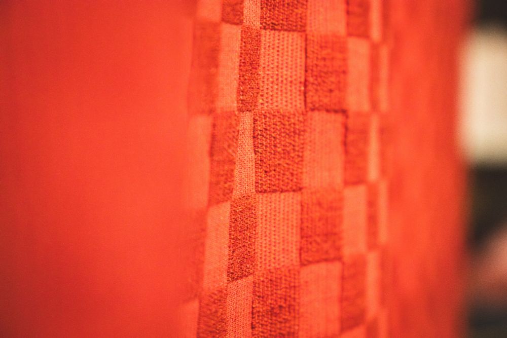 Das Bild zeigt eine Nahaufnahme eines vertikal drapierten, leuchtend orange-roten Stoffs mit einem karierten Muster (Schachbrettmuster).
​Vordergrund (Links): Die linke Seite des Bildes ist unscharf (geringe Schärfentiefe) und wird von einer großen, ungemusterten Fläche in einem satten, tiefen Orangerot dominiert.
​Muster (Mitte und Rechts): Der rechte Teil des Bildes ist scharf fokussiert und zeigt deutlich das karierte Muster. Es besteht aus abwechselnden Quadraten von zwei leicht unterschiedlichen Rottönen. Der Stoff hat eine fühlbare, grobe Webstruktur, die die einzelnen Quadrate bildet.
​Tiefe und Beleuchtung: Die Fokussierung liegt auf der Textur und dem Muster der rechten Seite, während die linke Seite verschwommen ist, was dem Bild eine gewisse Tiefe verleiht. Die Beleuchtung ist weich, betont die Textur des Stoffs und lässt das Orange-Rot intensiv leuchten.
​Gesamteindruck: Es handelt sich um eine abstrakte, texturierte Aufnahme, die sich auf Farbe, Stofflichkeit und Muster konzentriert.