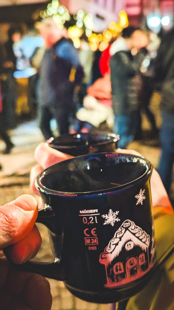Nahaufnahme einer dunkelblauen Glühweintasse mit Wintermotiven, die vor der unscharfen, beleuchteten Kulisse eines Weihnachtsmarktes bei Nacht in der Hand gehalten wird.