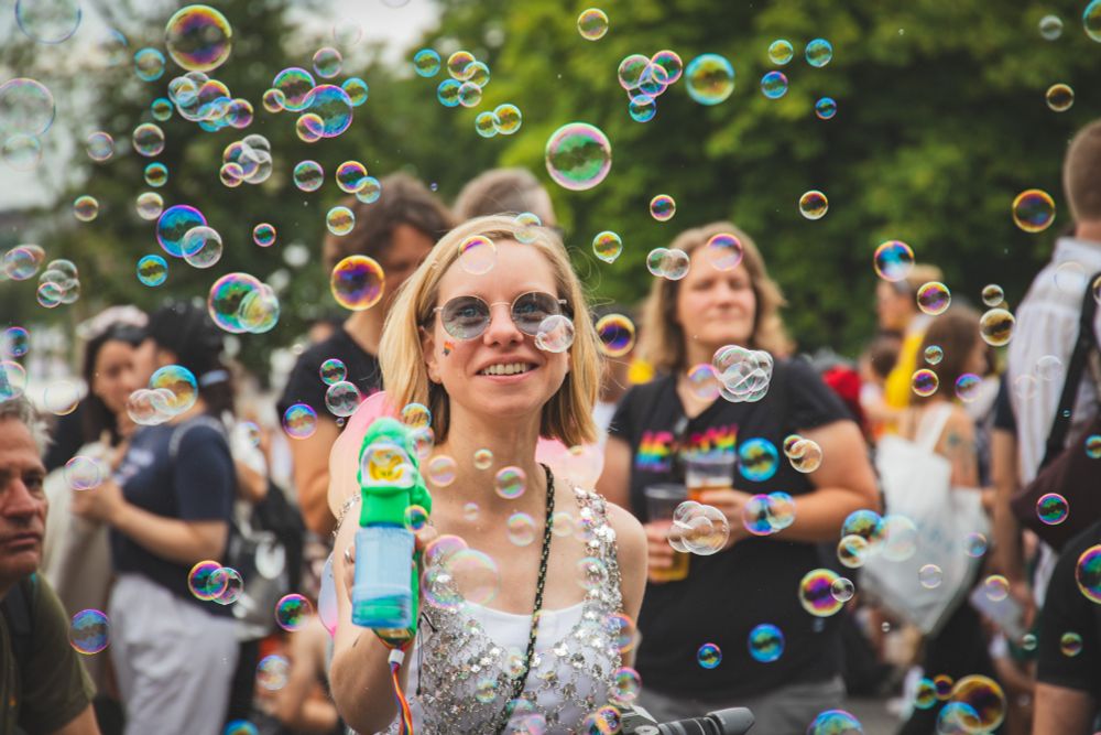 Das Bild zeigt eine junge, lächelnde Frau in der Mitte einer Menschenmenge im Freien, wahrscheinlich bei einer Veranstaltung oder Parade. Sie ist von zahlreichen, schillernden Seifenblasen umgeben, die den Vordergrund und die Mitte des Bildes dominieren.

Vordergrund und Zentrum: Die Frau hat kurzes blondes Haar, trägt eine weiße, glitzernde Trägershirt oder Oberteil und eine Sonnenbrille mit runden Gläsern. Sie hält in ihrer rechten Hand einen blauen Spielzeug-Seifenblasen-Puster in Form einer Comicfigur, mit dem sie die Blasen erzeugt. Auf ihrer Wange ist ein kleines, buntes Regenbogen-Tattoo oder -Aufkleber zu sehen. Sie blickt direkt in die Kamera und lächelt.

Hintergrund: Hinter ihr ist eine unschärfere Gruppe von Menschen zu erkennen. Eine weitere Frau mit blonden Haaren und einem schwarzen T-Shirt hält ein Glas oder einen Becher in der Hand und schaut ebenfalls lächelnd in die Menge. Links und rechts sind weitere Gesichter von Teilnehmenden sichtbar.

Atmosphäre: Die Beleuchtung ist hell, was auf einen sonnigen Tag hindeutet. Im obersten Hintergrund sind grüne Baumkronen sichtbar. Die allgemeine Stimmung des Bildes ist fröhlich, ausgelassen und festlich. Die vielen bunten Seifenblasen verleihen der Szene eine magische und unbeschwerte Atmosphäre.
