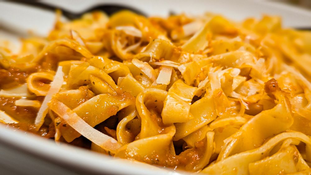 Eine appetitliche Nahaufnahme von breiten Bandnudeln, die glänzend mit einer roten Soße überzogen sind. Das Gericht ist großzügig mit groben Spänen von hellem Hartkäse, wie Parmesan, bestreut. Der Fokus liegt auf der Mitte des Gerichts, während der Hintergrund leicht verschwommen ist