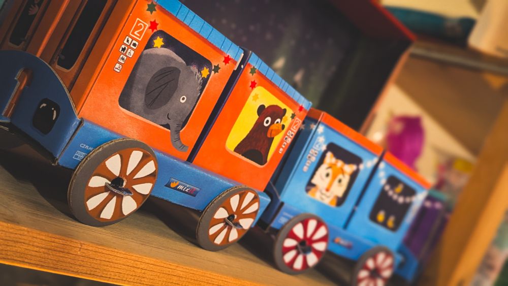 Fokus: Eine Nahaufnahme und seitliche Ansicht der mittleren und hinteren Waggons desselben bunten Spielzeugzugs aus Pappe.
​Die Waggons: Die Waggons sind in einem lebhaften Wechsel von Orange und Blau gehalten. Jeder Waggon hat ein großes, rechteckiges Fenster, hinter dem sich eine Illustration eines Tieres befindet, das das "Türchen" darstellt.
​Der vorderste sichtbare Waggon (Orange) zeigt einen grauen Elefanten mit der Nummer "2" in der oberen Ecke.
​Der nächste Waggon (Orange) zeigt einen braunen Bären.
​Der dritte sichtbare Waggon (Blau) zeigt einen weißen Tiger oder eine weiße Katze.
​Hinten folgt ein weiterer blauer Waggon, dessen Motiv dunkler ist und festliche, gelbe Lichterketten zeigt.
​Details: Die blauen Unterseiten der Waggons tragen das Logo "MIXC". Die Räder sind groß, rund und haben weiß-braune Speichenmuster.
​Umgebung: Der Zug steht auf einem Holzregal und das Bild ist warm beleuchtet, was die festliche Adventsstimmung unterstreicht.
​Fassen wir zusammen: Die Bilder zeigen einen detaillierten, weihnachtlichen Adventskalender in Form eines Karton-Spielzeugzugs, dessen Waggons bunte Illustrationen von Lokführern, Engeln und Tieren (wie Elefant, Bär und Tiger) zeigen, welche die einzelnen Kalendertürchen darstellen.