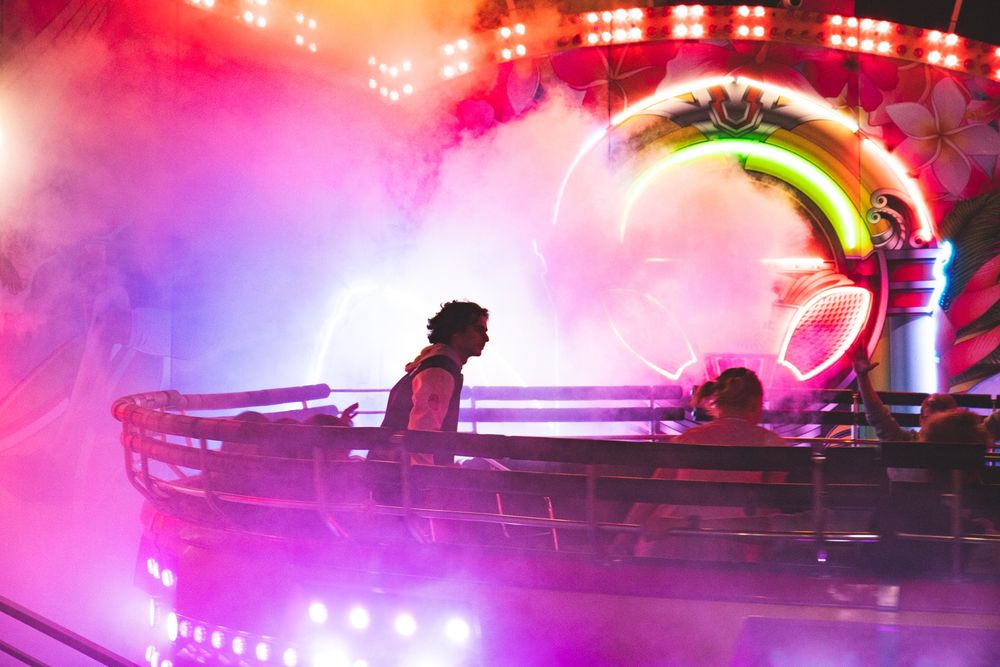 Dies ist eine dynamische und farbenfrohe Nachtaufnahme, die wahrscheinlich ein Jahrmarkt- oder Kirmes-Fahrgeschäft zeigt, möglicherweise ein Karussell oder eine Art sich drehende Plattform, die von grellem, künstlichem Licht und Nebel oder Rauch eingehüllt ist.

Beleuchtung und Atmosphäre
Das Bild wird dominiert von intensiven, leuchtenden Farben, insbesondere leuchtendem Pink, Lila und hellem Rot.

Ein dichter rosa-weißer Rauch oder Nebel durchzieht die Szene und wird von den Lichtern angestrahlt, was eine dramatische, fast surreale Atmosphäre schafft.

Im Hintergrund ist die Neon-Beleuchtung des Fahrgeschäfts selbst zu sehen, die helle, bogenförmige oder kreisförmige Formen in Grün, Gelb und Rot zeigt.

Personen
Im mittleren Teil des Bildes ist eine Gruppe von Menschen zu sehen, die in einer runden oder bootförmigen Gondel des Fahrgeschäfts sitzen oder stehen.

Eine zentrale Figur steht seitlich in der Gondel, mit dunklem, lockigem Haar, und blickt nach rechts. Die Silhouette des Körpers ist scharf gegen das helle Pink und Lila im Hintergrund abgehoben.

Weitere Personen sind rechts im Schatten oder von hinten zu sehen, wobei eine Person ihren Arm nach oben gestreckt hält.

Fahrgeschäft
Der sichtbare Teil des Fahrgeschäfts ist die dunkle, metallische Umrandung der Gondel, die sich horizontal durch das Bild zieht.

Helle weiße Lichtakzente von unten leuchten die Gondel und den Nebel an.

Zusammenfassend lässt sich sagen: Es ist eine lebendige Szene voller Energie und Bewegung, die die aufregende und neongrelle Atmosphäre eines nächtlichen Jahrmarkts einfängt.
