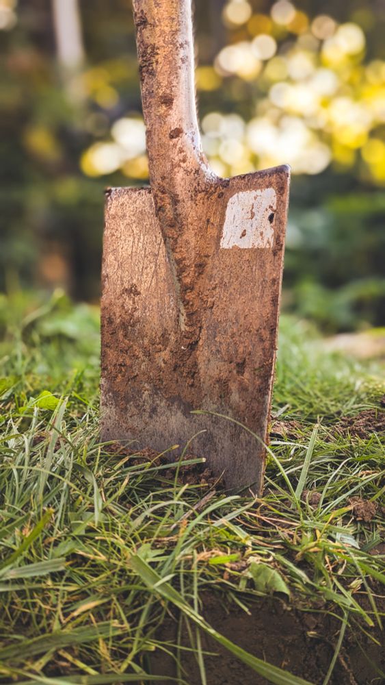 Eine Nahaufnahme des Blattes einer Gartenschaufel oder eines Spatens, das aufrecht im Gras steckt. Das Metall des Schaufelblatts ist mit trockener Erde und Schmutz bedeckt. Auf dem Schaufelblatt ist ein kleiner, rechteckiger, heller Aufkleber oder eine Markierung sichtbar. Die Schaufel steckt in kurzem grünen Gras und Erde. Im unscharfen Hintergrund sind verschwommene, helle, gelblich-grüne Lichter zu sehen, die auf einen sonnigen Tag in der Natur oder im Garten hinweisen.