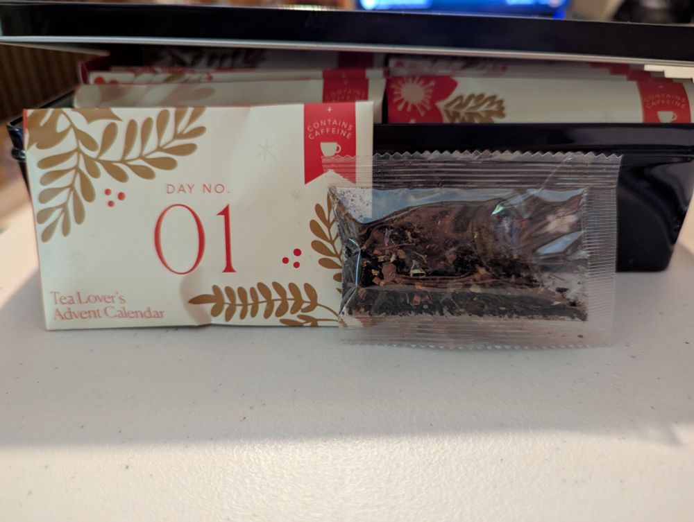 The envelope for day 1 of the tea calendar with the clear pouch containing the loose leaf tea next to it. The envelope is white with red "DAY NO. 01" in the center, gold branches/leaves at the corners, and "Tea Lover's Advent Calendar" in the bottom left corner. It is a caffeinated selection of teas.