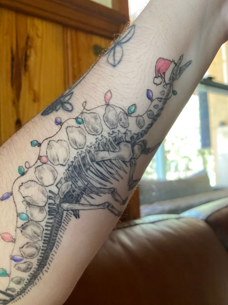 A fine line tattoo of a stegosaurus skeleton with a Santa hat and Christmas lights drawn on in ballpoint pen. 