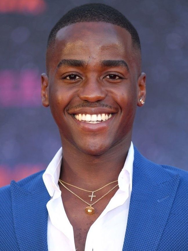 Ncuti Gatwa wearing a blue jacket and gold necklaces and earrings facing the camera directly