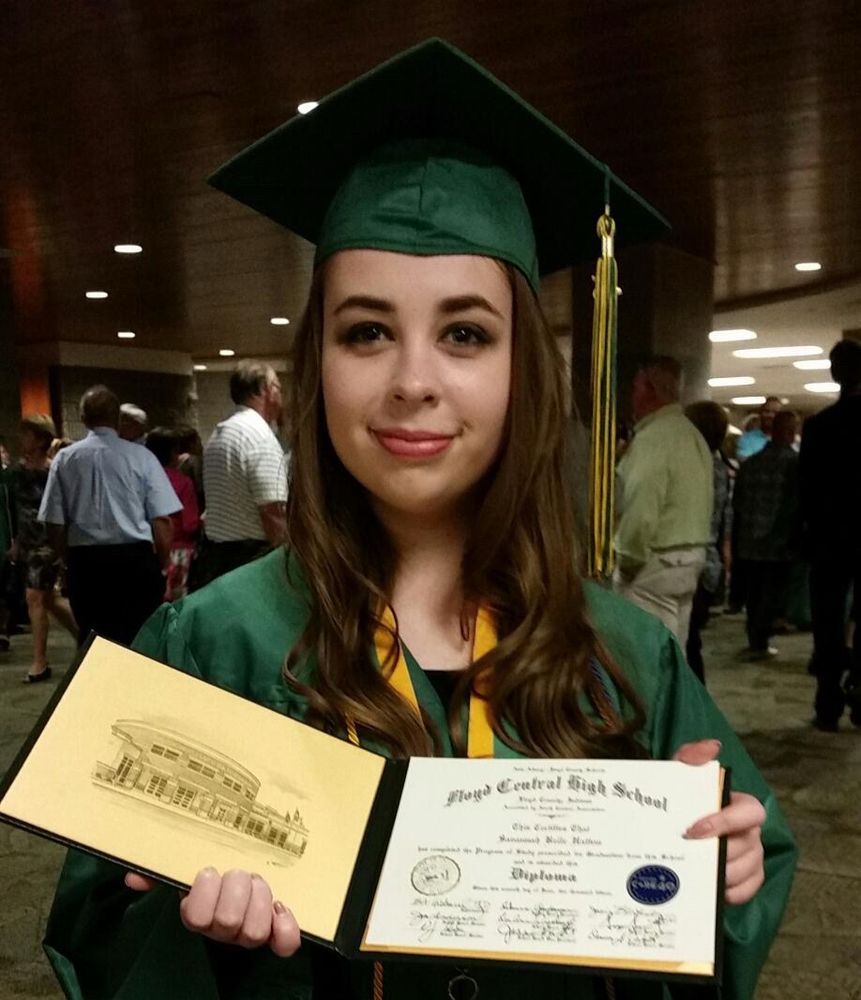 19 year old Vannah, long brown hair, natural eyeshadow stands in a Green cap and gown holding their high school diploma 