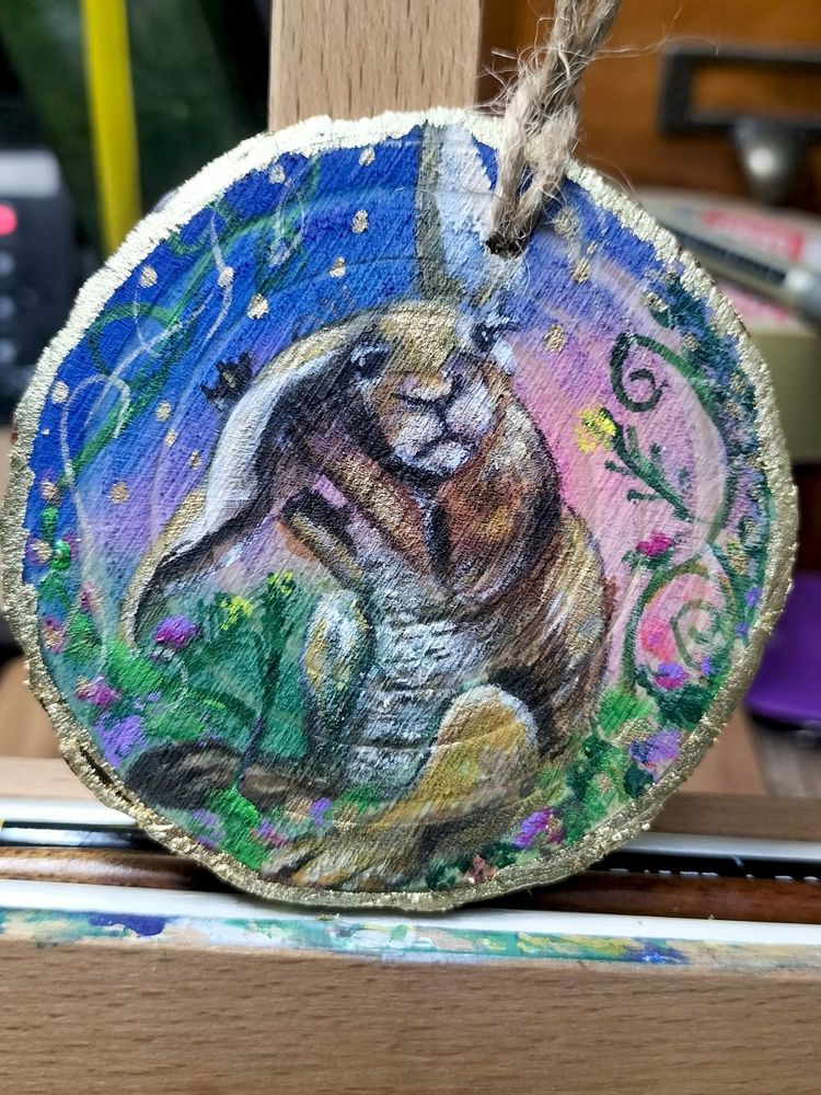 Whimsical small wood slice painting of a sitting hare, rubbing his ear.  Background is soft pink and dark blue, with colourful flowers and swirls and gold highlights.   Lovely Spring depiction 