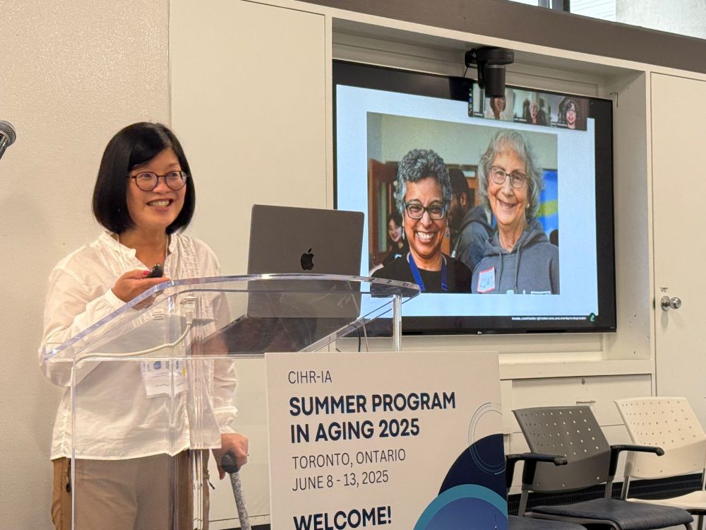 Dr. Lillian Hung, Canada Research Chair in Senior Care, speaks at a podium during the CIHR Institute of Aging Summer Program in Aging 2025 in Toronto. A large screen behind her displays a slide featuring two smiling family partners and co-researchers, Bubli Chakraborty and Lynn Jackson. 