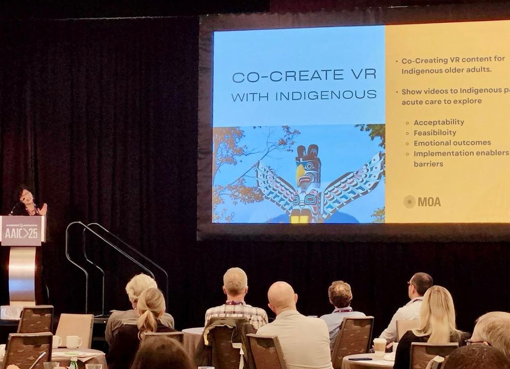 Dr. Lillian Hung speaking on stage at a podium labeled “AAIC 25,” with a large presentation slide behind her titled “Co-Create VR with Indigenous.” The slide includes a photo of a totem pole and a bulleted list outlining the goals of co-creating VR content with Indigenous older adults.