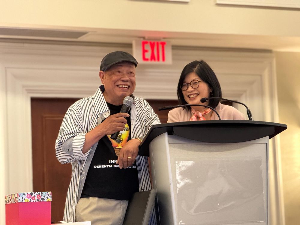 Mario Gregorio, a smiling older man wearing a cap and striped shirt, stands at a podium holding a microphone. Beside him is Dr. Lillian Hung, also smiling, in a pink blazer and glasses. They are co-presenting at the 2025 West Coast Conference on Aging.