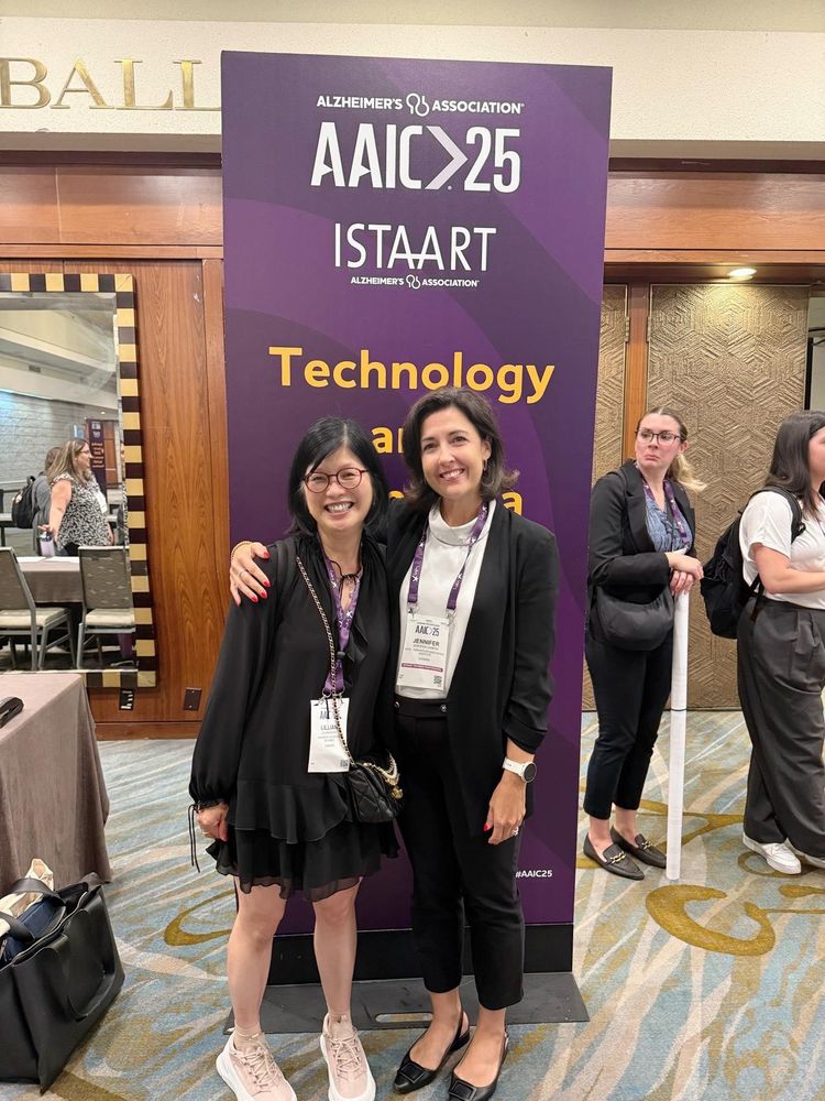 Dr. Lillian Hung and Dr. Jennifer Campos smiling and posing together in front of a tall purple sign that reads “AAIC 25, ISTAART, Technology and Dementia” at the Alzheimer’s Association International Conference 2025 preconference venue.