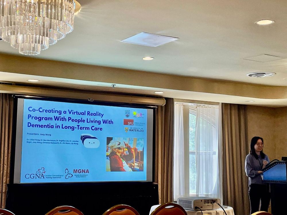 Joey Wong stands at a podium presenting a slide titled “Co-Creating a Virtual Reality Program With People Living With Dementia in Long-Term Care.” The slide shows logos from UBC, SFU, the University of Waterloo, CGNA, and the MGNA, and includes a photo of an older adult wearing a VR headset.