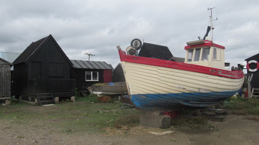 Rough ground, cloudy grey sky, black sheds. In front of them, a small battered boat on blocks. It is painted white with red and blue trim. On the shed on the far right of the frame, a life belt with the label 'MOJO'.