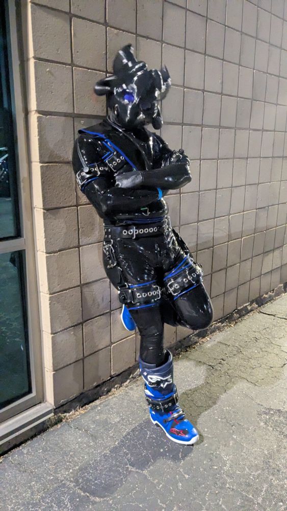 Fully covered rubber drone leaning against a wall wearing a custom rubber hood, rubber restraints, and motocross boots.