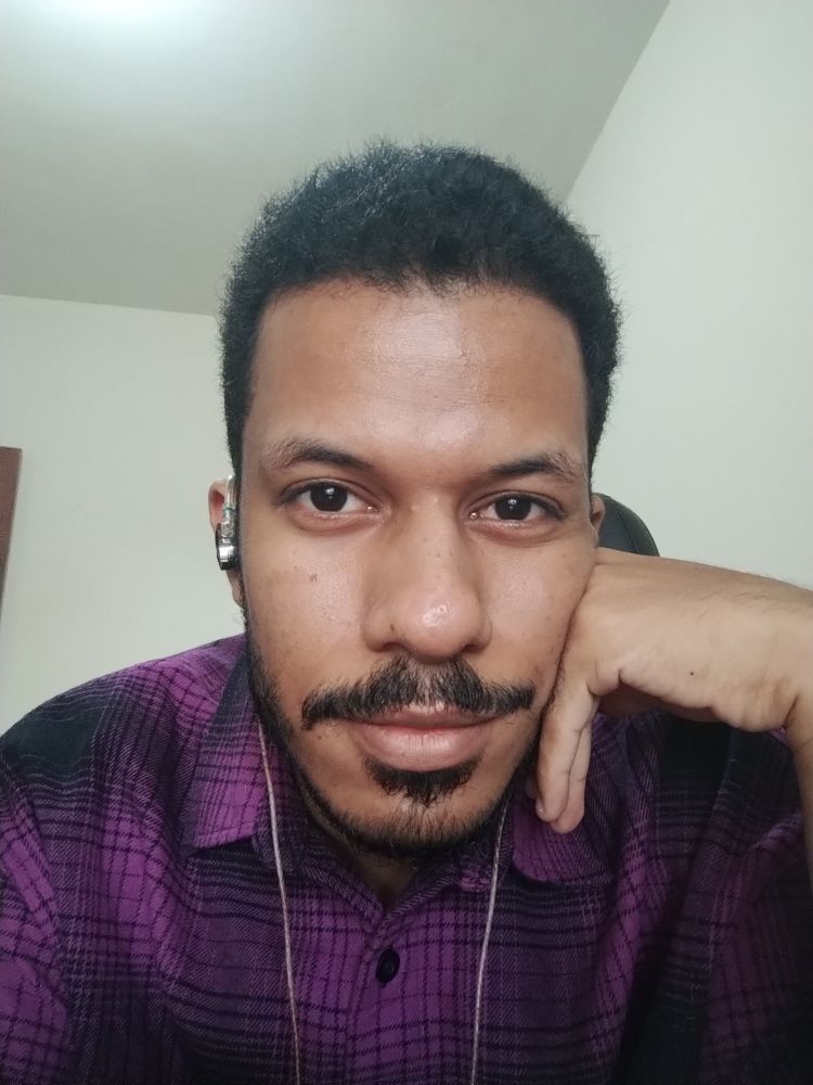 Foto do rosto de um homem negro bem perto com um pouco de bigode e barba pretos, cabelo preto também. O homem está vestindo uma camisa roxa de botão.