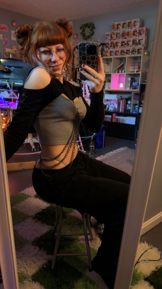 clover sitting on a stool informs of a mirror taking a selfie in the mirror. they’re sitting sideways and smiling while winking at the camera. 