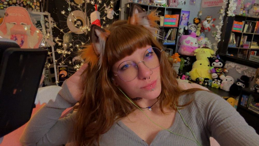 clover taking a selfie at their desk. they’re looking at the camera and smiling slightly. they have fox ears on. 