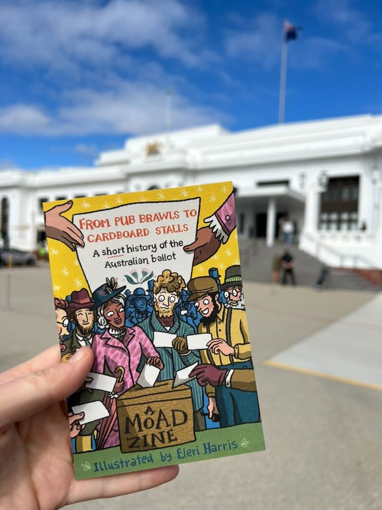 Hand holding mini comic in front of old parliament house in Canberra, Australia. The comic has lots of people in historic costume on the cover holding ballot slips behind a ballot box and says "From pub brawls to cardboard stalls: A short history of the Australian Ballot, a MoAD zine illustrated by Eleri Harris" 