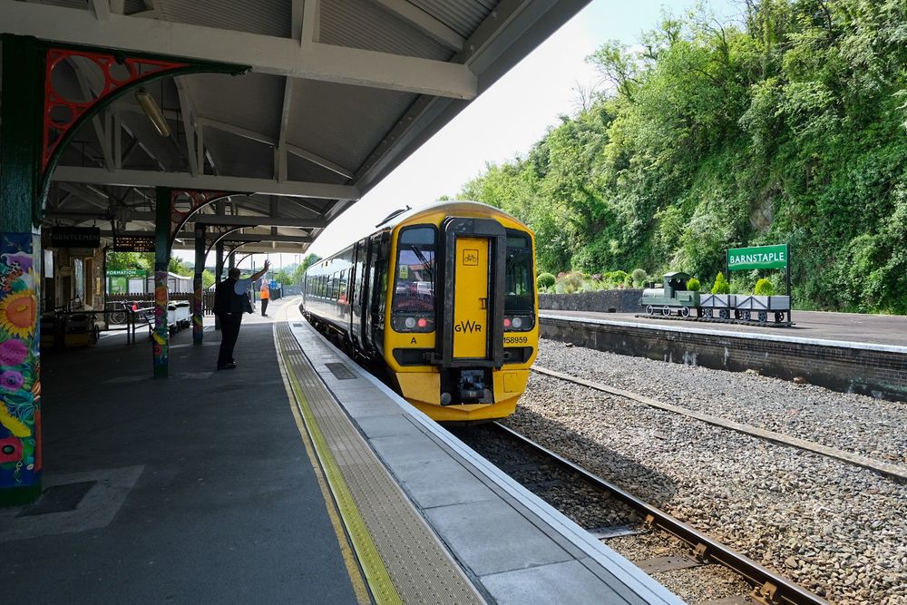 A GWR train waits at Barnstaple station. It is a sunny day and a member of GWR staff waves at the train from the platform