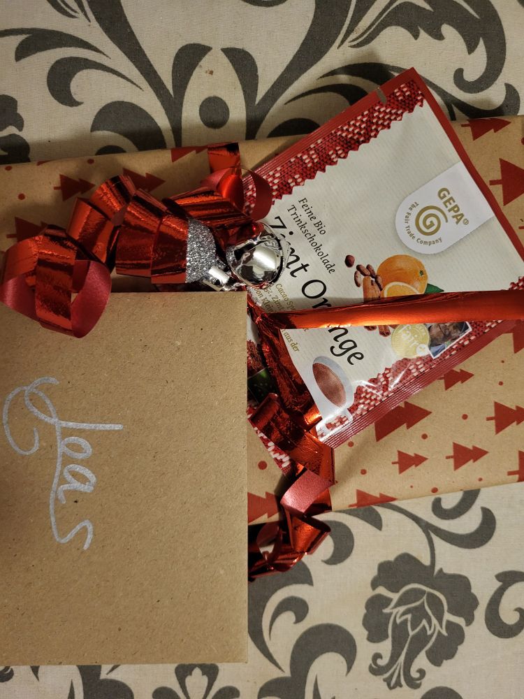 Ein in weihnachtlichem Papier verpacktes Geschenk, eine Tüte "Feine Bio Trinkschokolade Zimt Orange", eine Karte, auf der das Wort "Joa" steht.