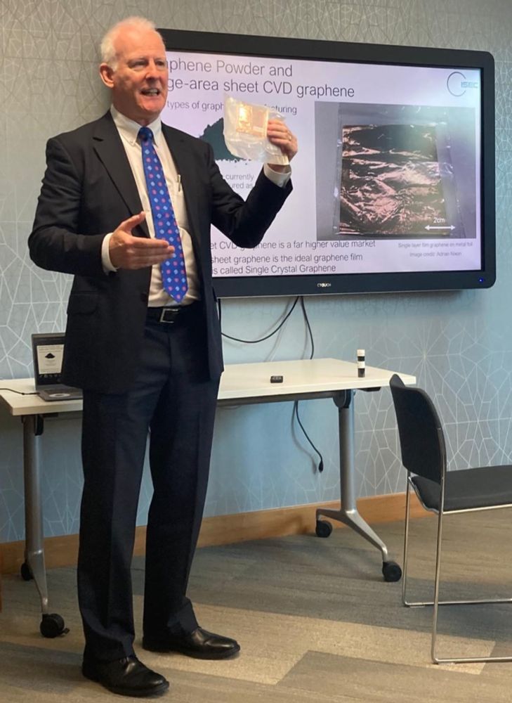 Adrian Nixon presenting at a conference showing the difference between CVD graphene (held in his hand) and graphene powder in the vial on the table behind him with a slide in the background showing the difference between the to forms of the material