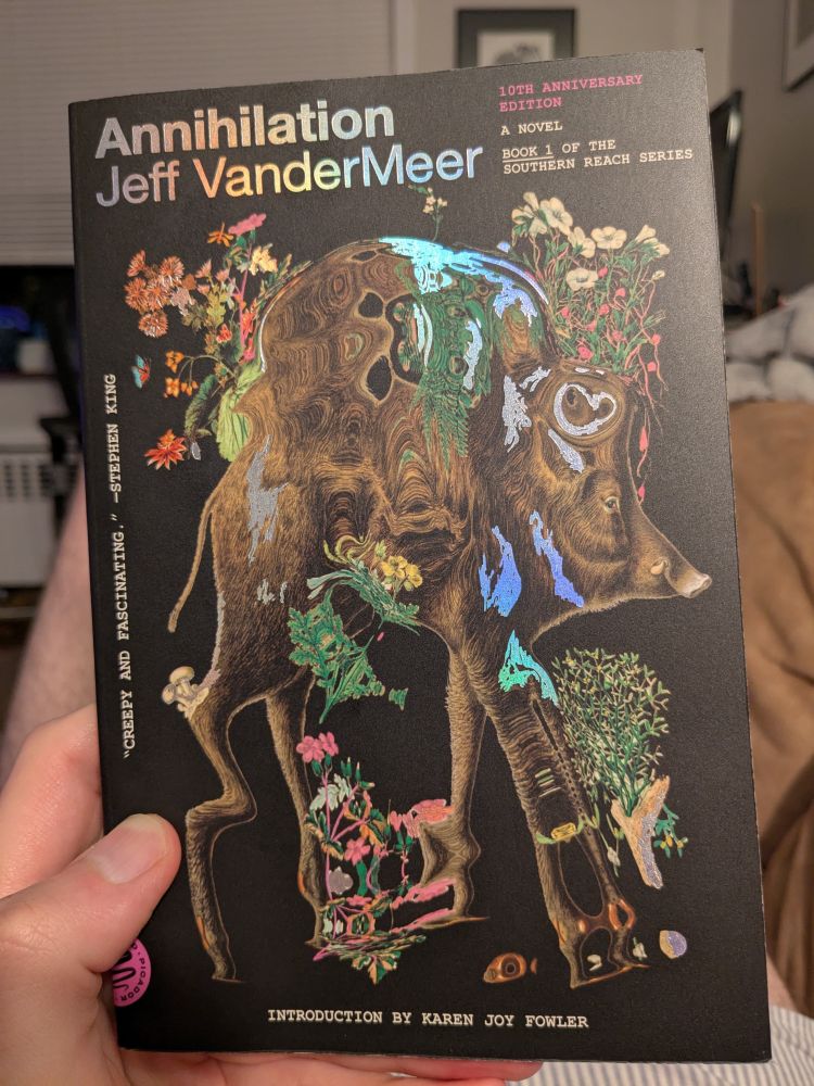 A hand holding a paperback copy of Annihilation by Jeff VanderMeer. The cover is illustrated with a surrealist painting of a boar with botanical drawings.