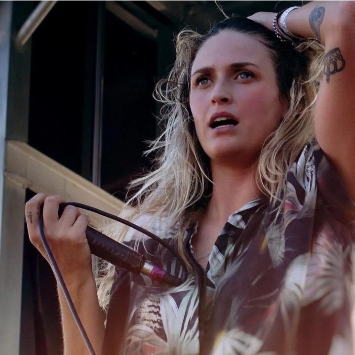 Emily Armstrong com camisa florida de fundo escuro, segurando o cabelo com uma das mãos e o microfone com outra.