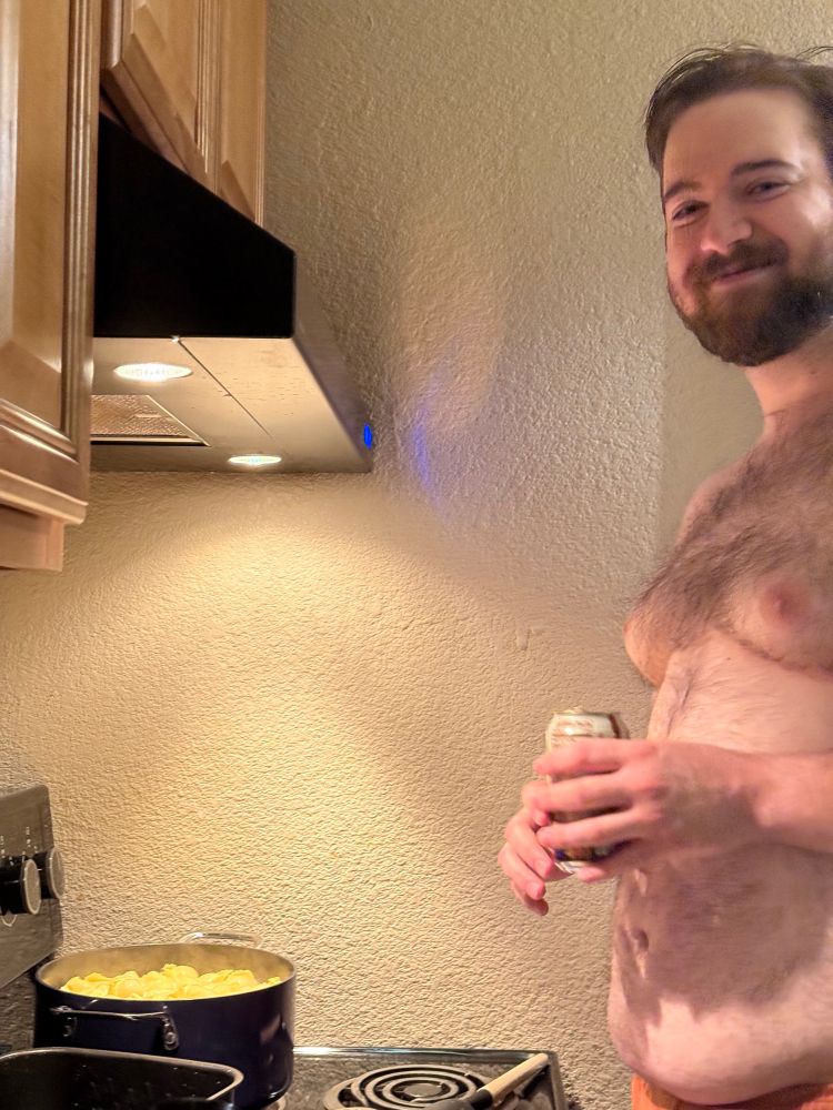 A shirtless man smiles at the camera holding a can of root beer in front of a stovetop over a pot of pasta