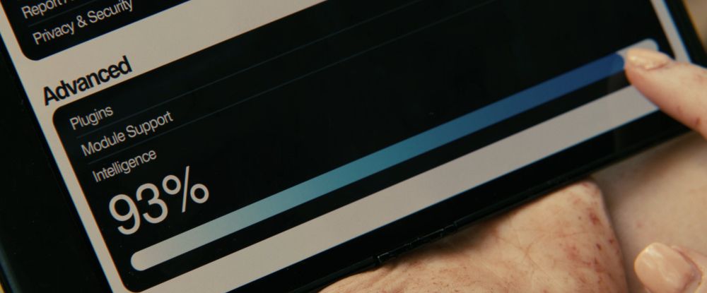 Film still depicting a person holding a tablet and adjusting an "Intelligence" slider on the screen. The slider is set to 93%.