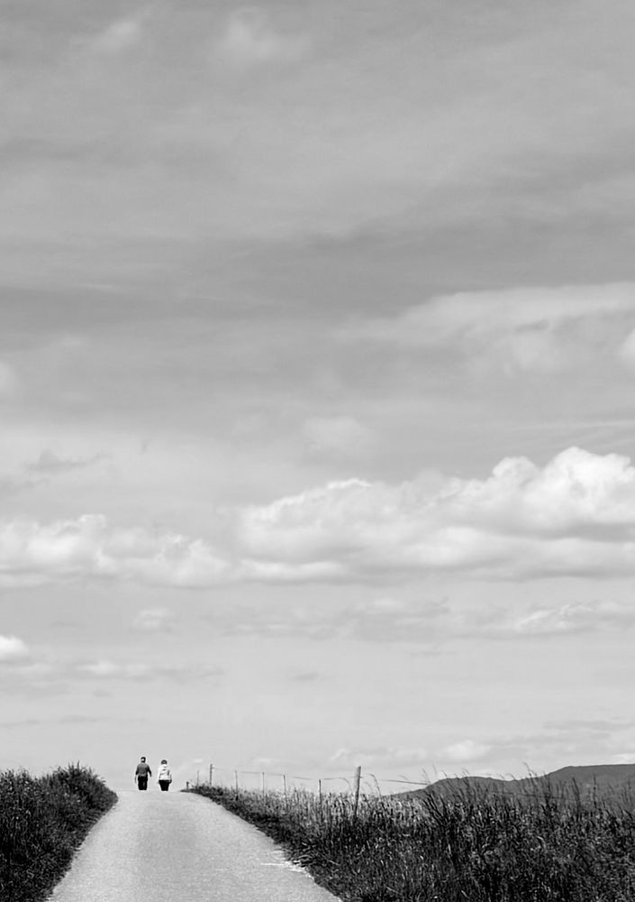 Black and white photo: Two small figures walking down a long, empty country road, gradually vanishing into the vast horizon beneath a wide, cloud-filled sky - as if they’re about to fall off the edge of the world.