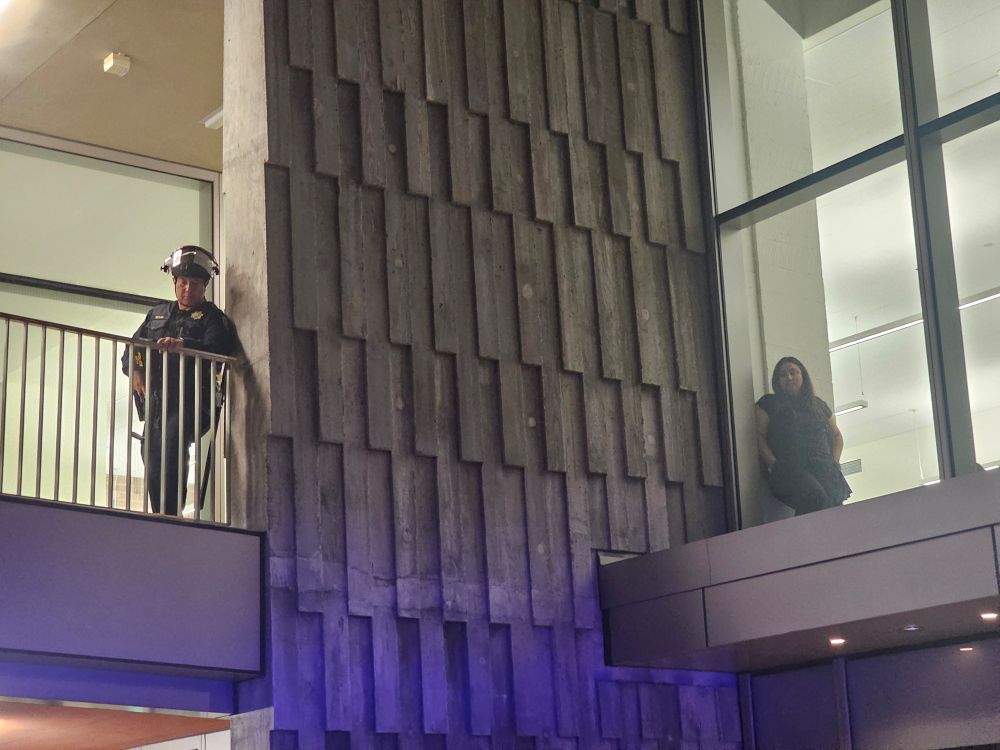 An officer wearing a helmet rests against the wall above the protesters. To the right, inside the student store, a worker watches the protest below. 
