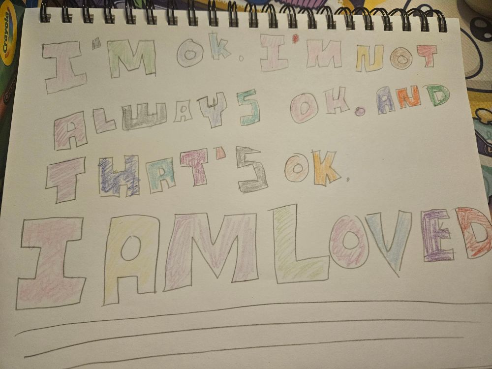 Poorly drawn art in a sketchbook that reads "I'm ok. I'm not always ok. And that's ok. I AM LOVED" it is colored in no particular pattern but many colors. 
