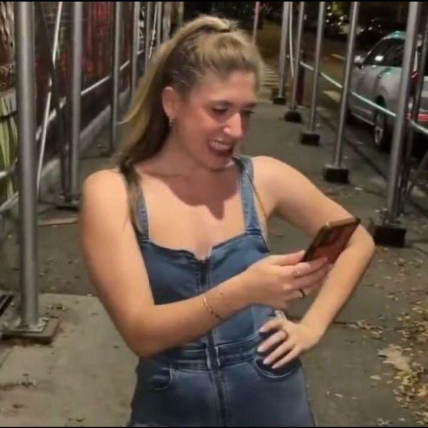 Uma mona loira usando uma jardineira jeans com o braço esquerdo apoiado na cintura e o direito segurando um celular, a qual ela encara com um sorriso amarelo e uma cara de "putz..."