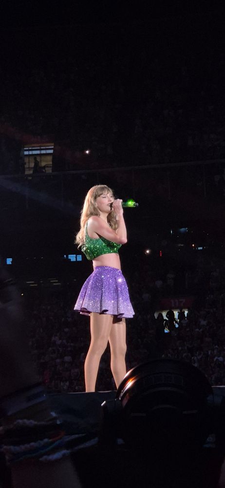 Taylor swift at the eras tour holding up a green mic during the 1989 set, wearing a green top and purple skirt