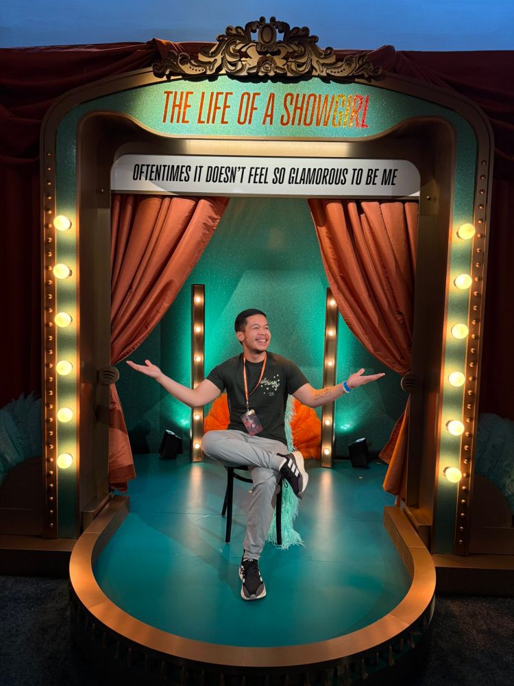 A broadway like stage. Its mostly square and at the front bexomes circle like. It has a gold trim. The back ia teal with orange curtains. There is a teal boarder that has orange lettering of "The Life Of A Showgirl". Under it there is a board with the saying "Oftentimes it doesnt feel so glamorous to me". At the front theres a chair that a man is sitting on. On the back of the chair theres an orange fan and teal boa