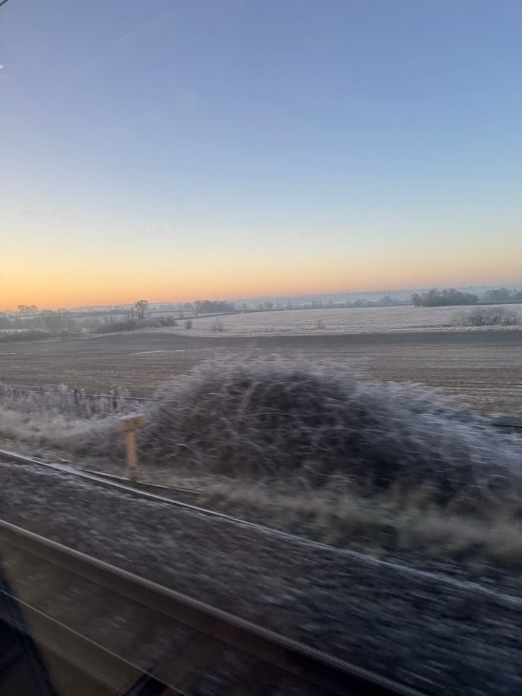 Frost-covered fields at sunrise, viewed from a moving train. The sky transitions from deep blue to warm orange near the horizon. Railway tracks run parallel in the foreground, with frost-coated grass on the embankment