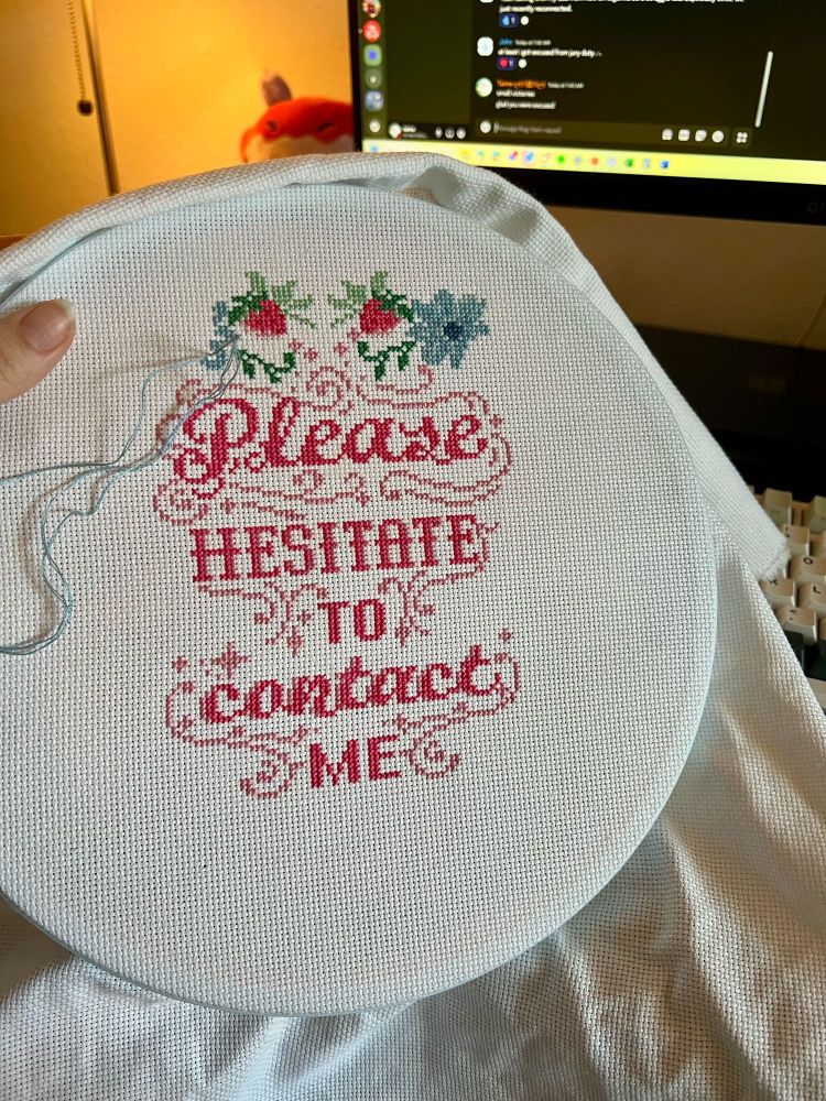 a photo of a cross stitch in progress that reads “please hesitate to contact me” written in a pink, stylized font